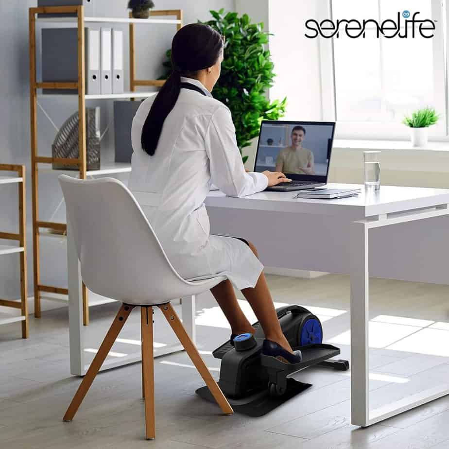 Lady exercising with the SereneLife SLEPL9 Under-Desk Elliptical Trainer while doing her desk work in her room.